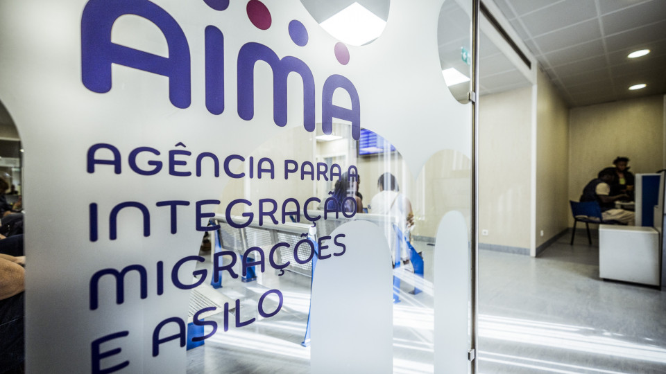Entrada de um escritório da Agência para a Integração, Migrações e Asilo (AIMA), com o nome da agência visível numa porta de vidro e pessoas a serem atendidas no interior.