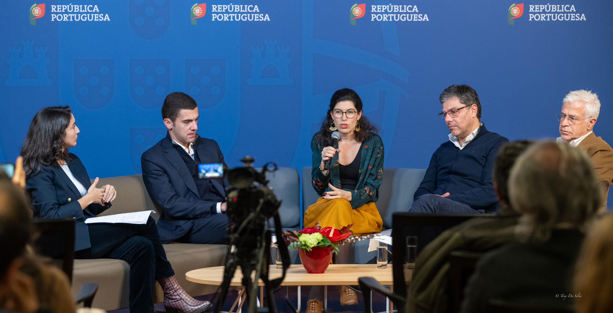 Painel de discussão com cinco pessoas sentadas em frente a uma plateia, num ambiente formal, com fundo azul.