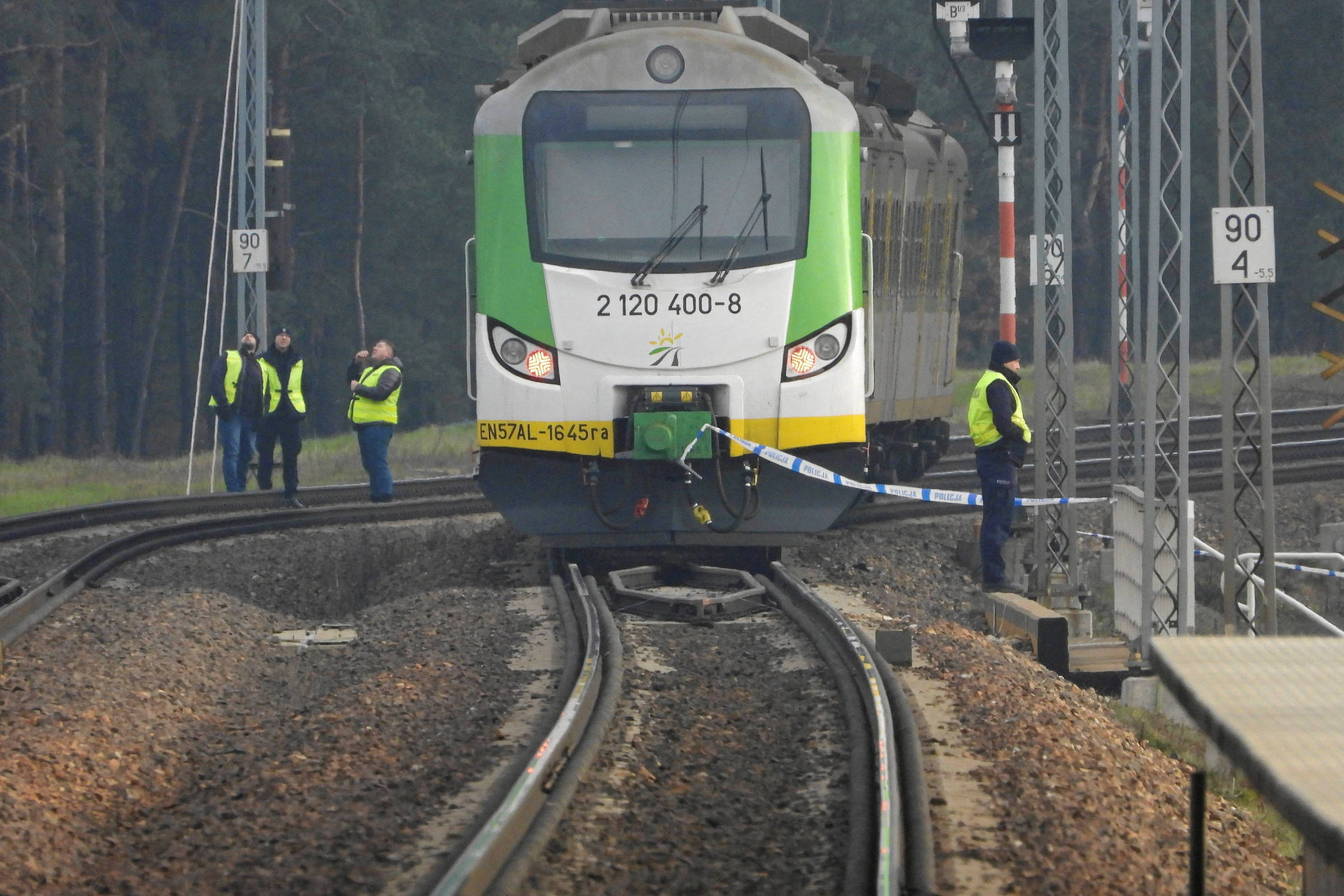 Comboio parado numa linha férrea danificada, rodeado por agentes de segurança, ilustrando a investigação de um ato de sabotagem ferroviária na Polónia.