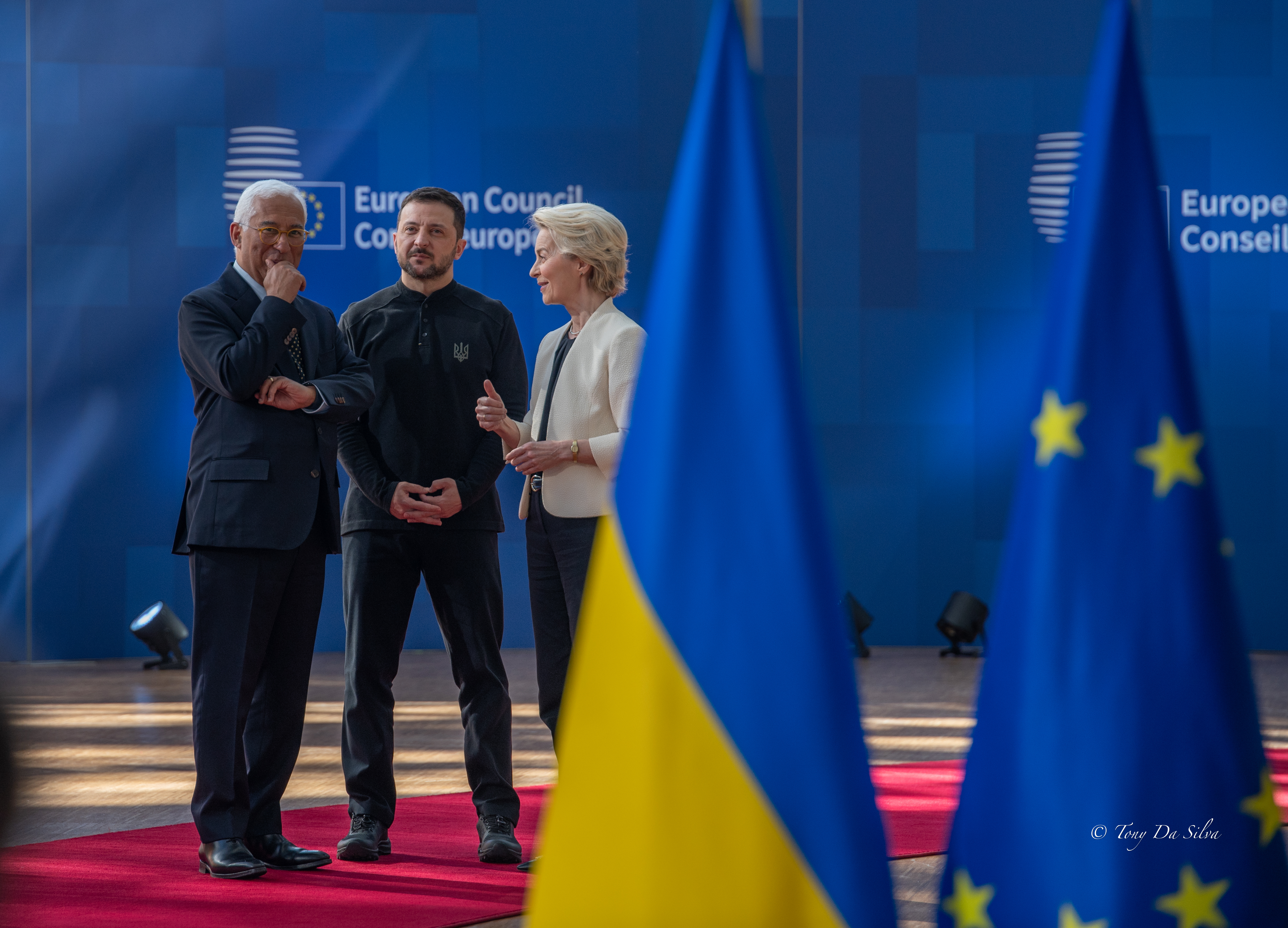 Três líderes políticos conversam num evento oficial, com bandeiras da União Europeia e da Ucrânia em destaque no primeiro plano, simbolizando negociações entre a UE e a Ucrânia.