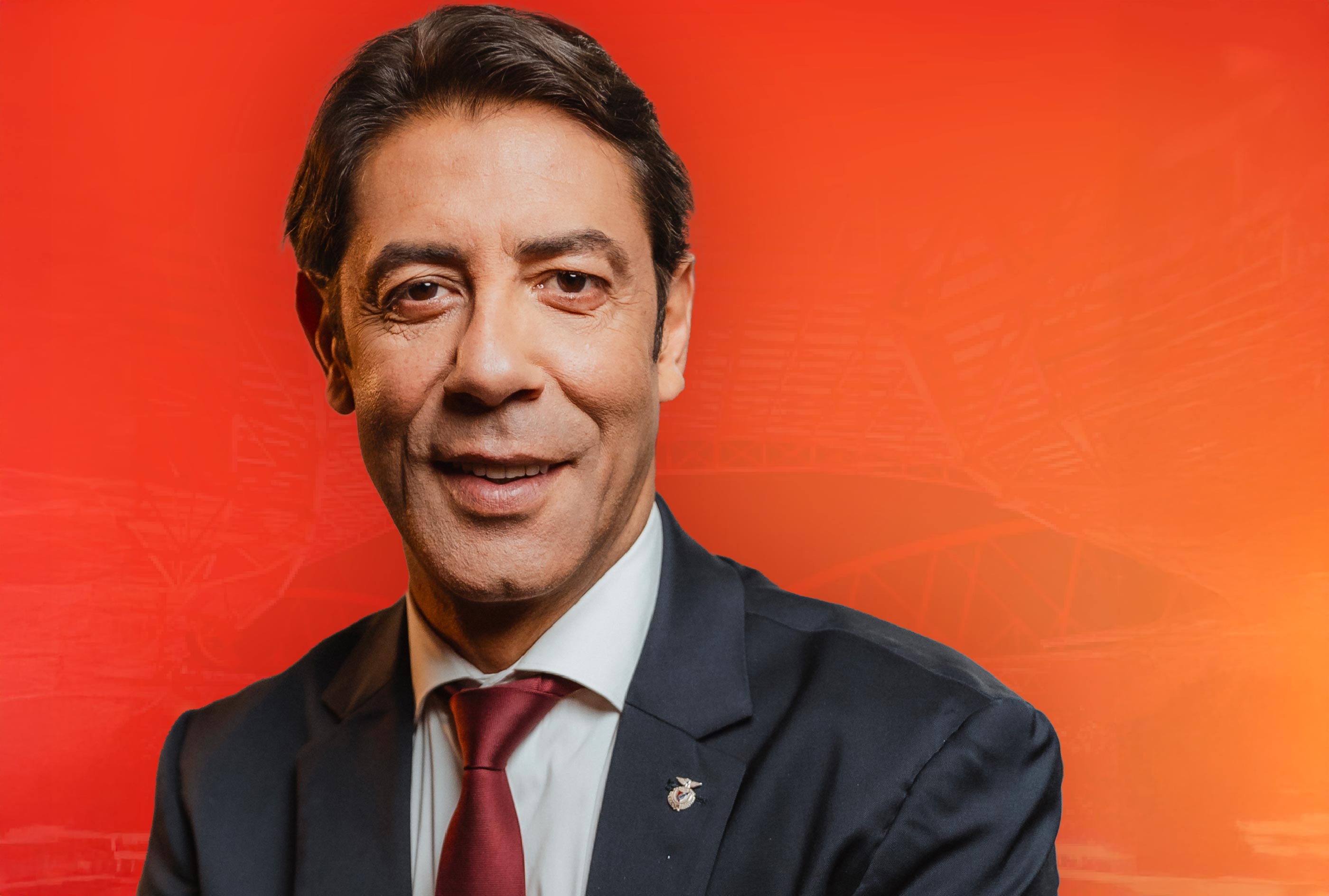Homem de fato escuro e gravata vermelha, sorridente, em frente a um fundo vermelho, representando liderança no contexto do Sport Lisboa e Benfica.