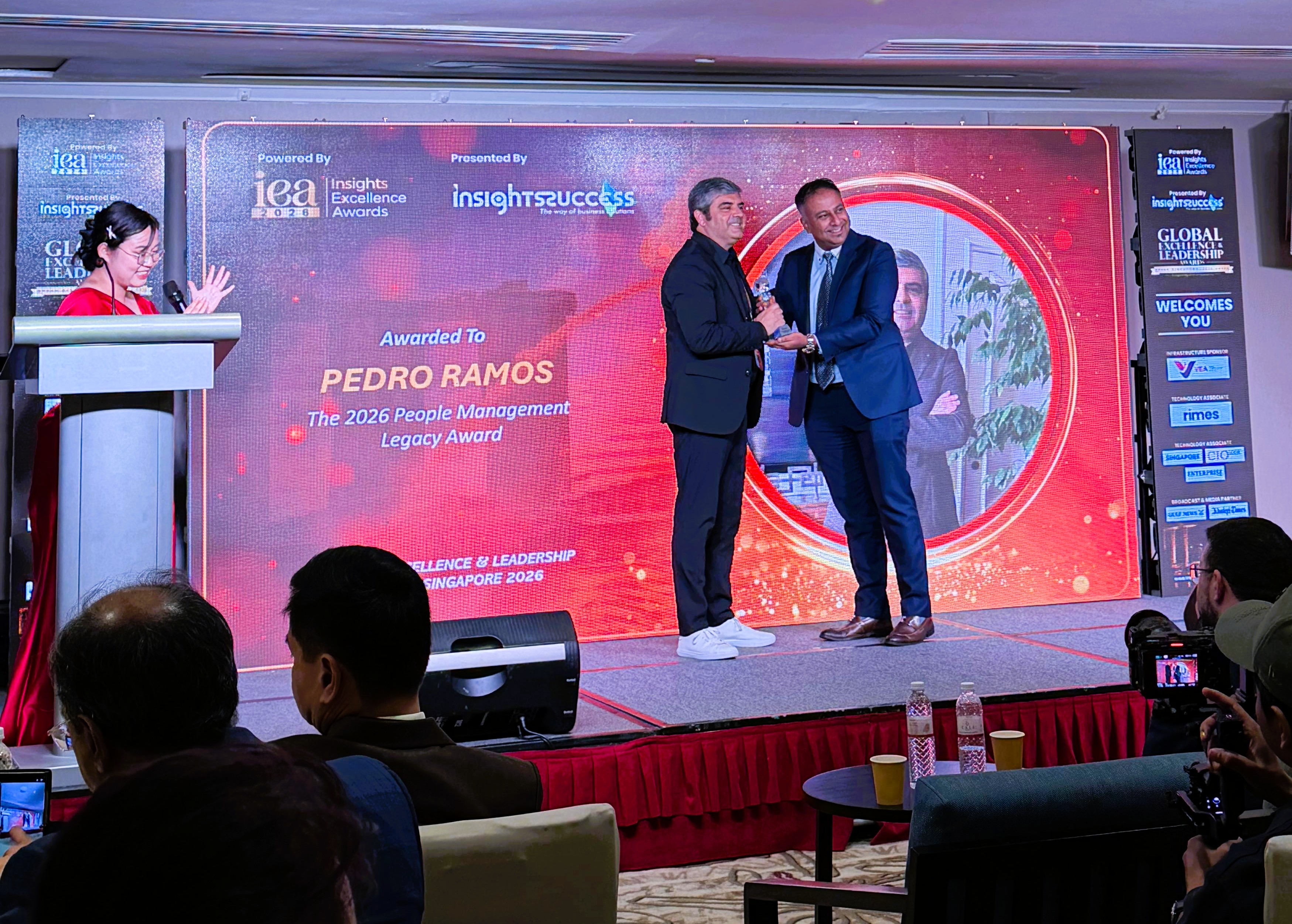 Dois homens em fato apertam as mãos no palco durante a cerimónia de entrega de prémios 'Global Excellence & Leadership Awards Singapore 2026', com uma apresentadora à esquerda e público sentado a assistir.