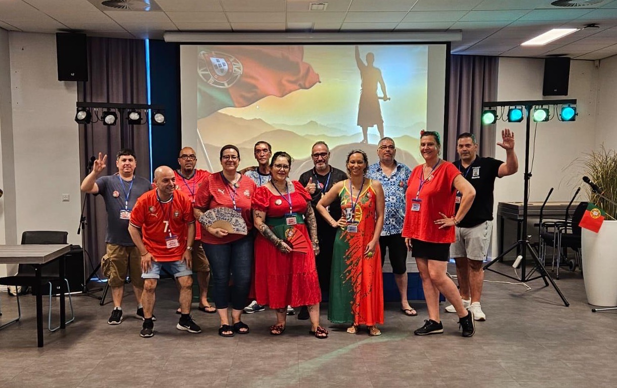 Grupo de pessoas reunido no centro de uma sala ampla, vestidas de forma colorida e festiva, em ambiente de convívio. Ao fundo, vê-se um ecrã com imagem projetada, reforçando o cenário de evento comunitário português em Tilburg.