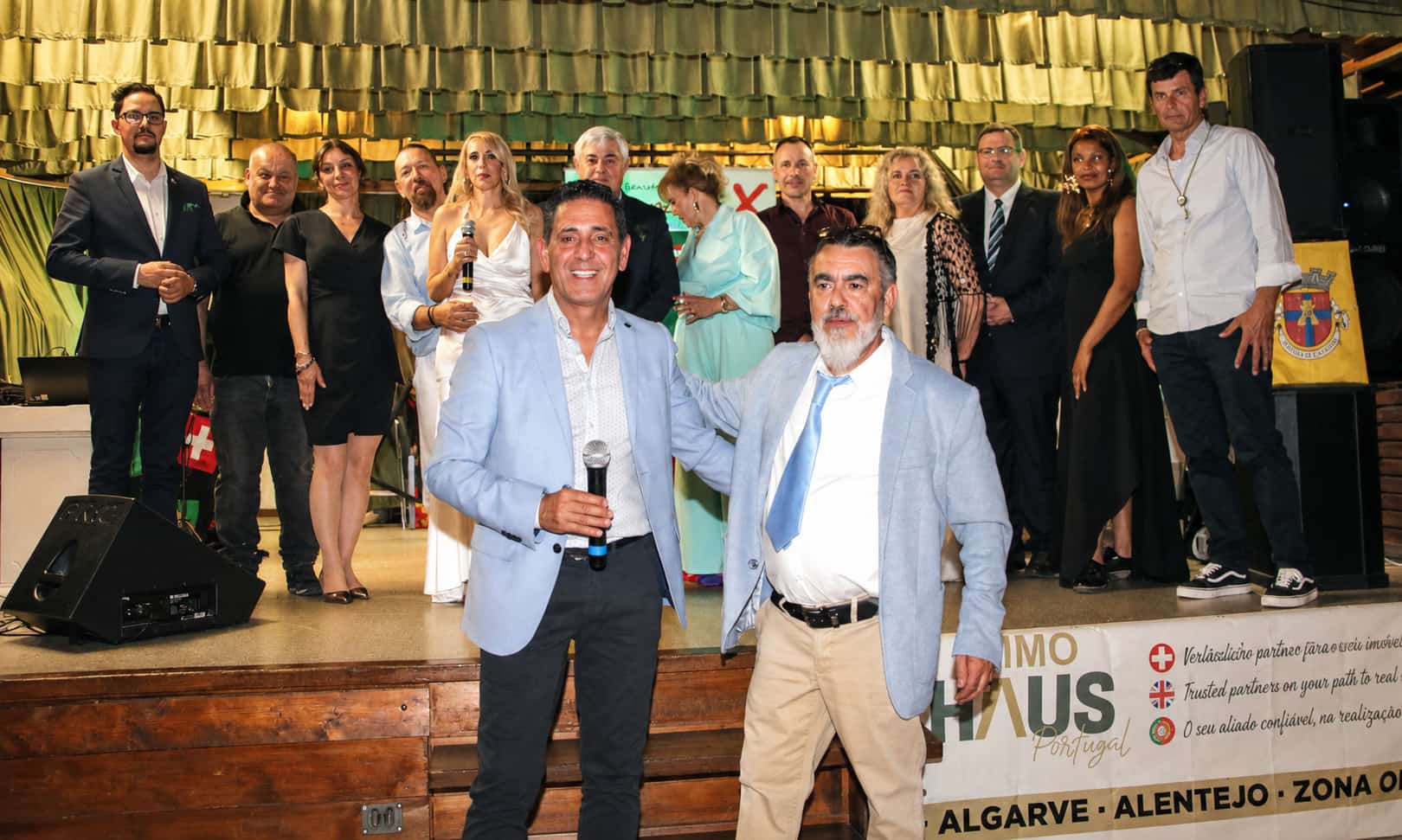 Grupo de pessoas em palco durante a 14.ª Gala Revista Repórter X, com dois indivíduos em destaque à frente, num ambiente de celebração e homenagem à cultura portuguesa.