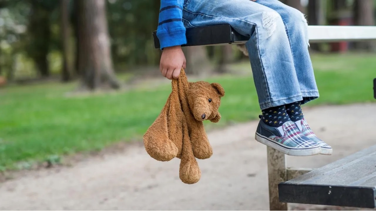 Criança sentada num banco de jardim, segurando um ursinho de peluche, sugerindo vulnerabilidade e inocência infantil.