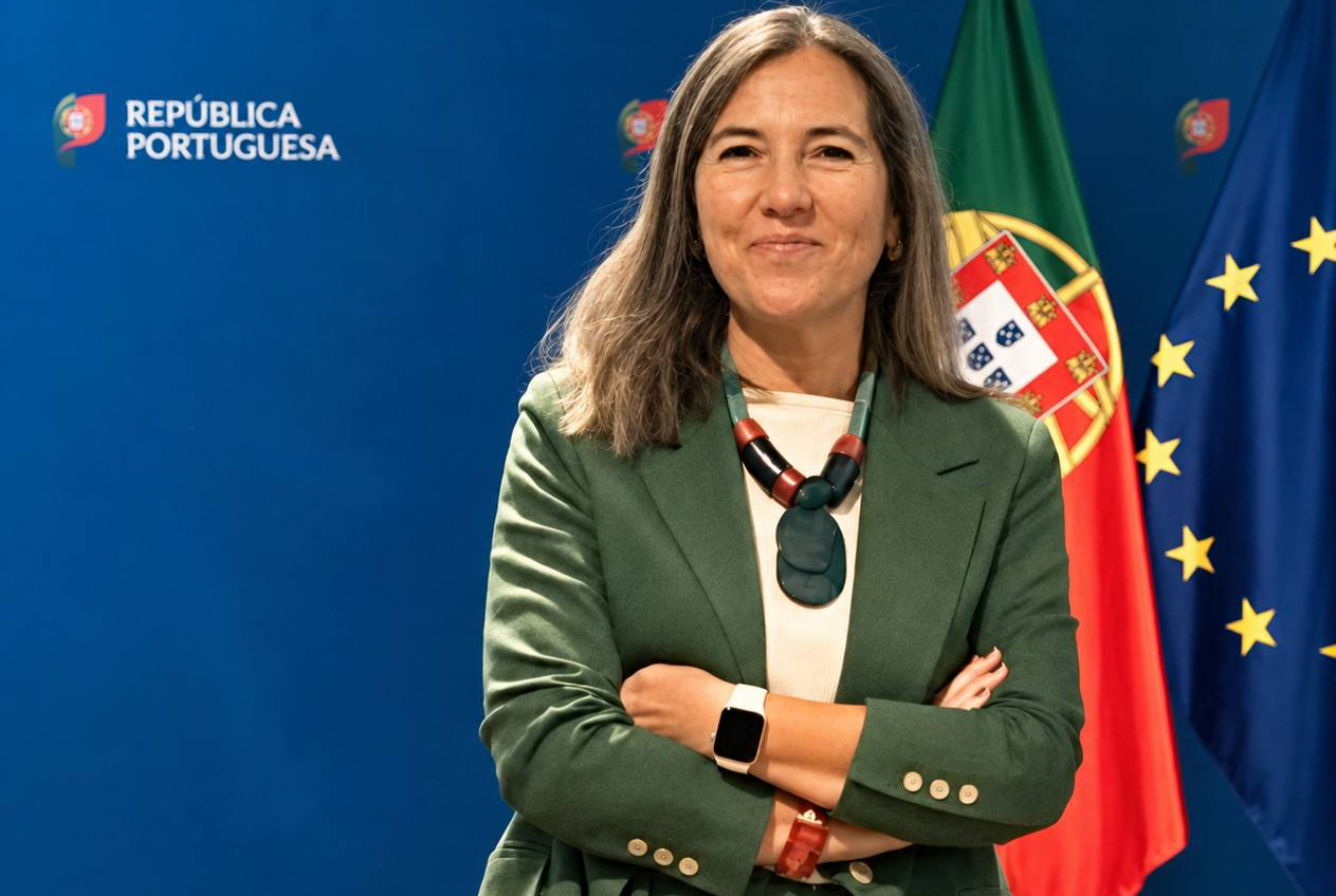Mulher de cabelo castanho, com blazer verde, de braços cruzados, em frente às bandeiras de Portugal e da União Europeia, num cenário institucional.