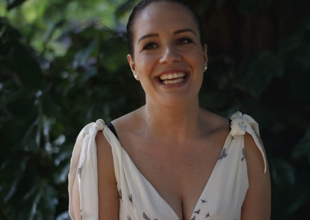 Mulher jovem de pele clara e cabelo escuro apanhado, sorridente, vestida de branco, fotografada num ambiente exterior com vegetação ao fundo. A imagem transmite alegria e vitalidade, em linha com a descrição de Suzy no artigo.