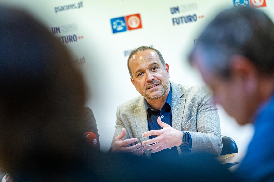 Francisco César, líder do PS/Açores, fala gesticulando numa reunião formal, com fundo decorado com logótipos e frases do partido, acompanhado por outras pessoas desfocadas em primeiro plano.