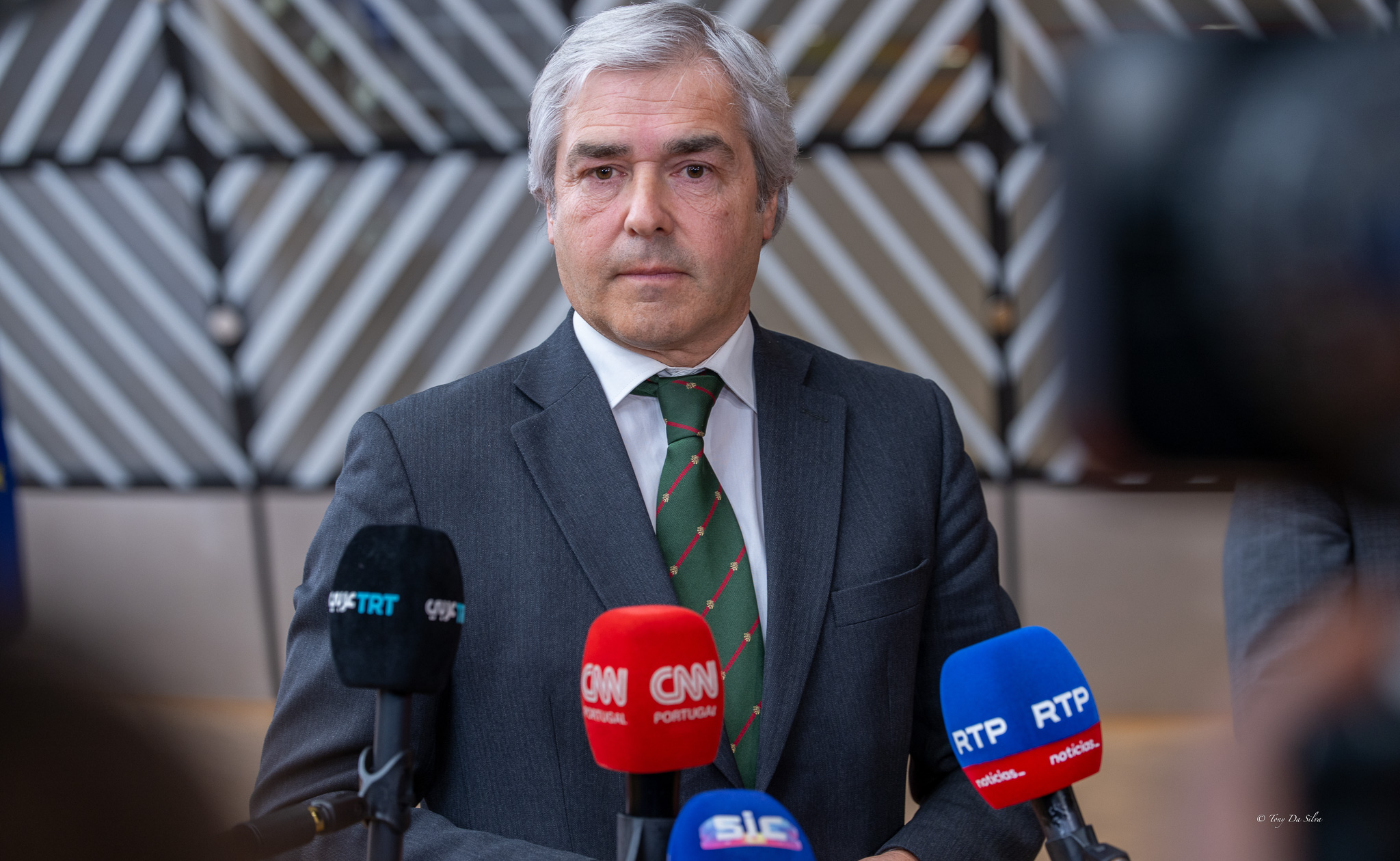 Ministro da Defesa português, de fato e gravata, fala numa conferência de imprensa perante microfones de vários meios de comunicação social.