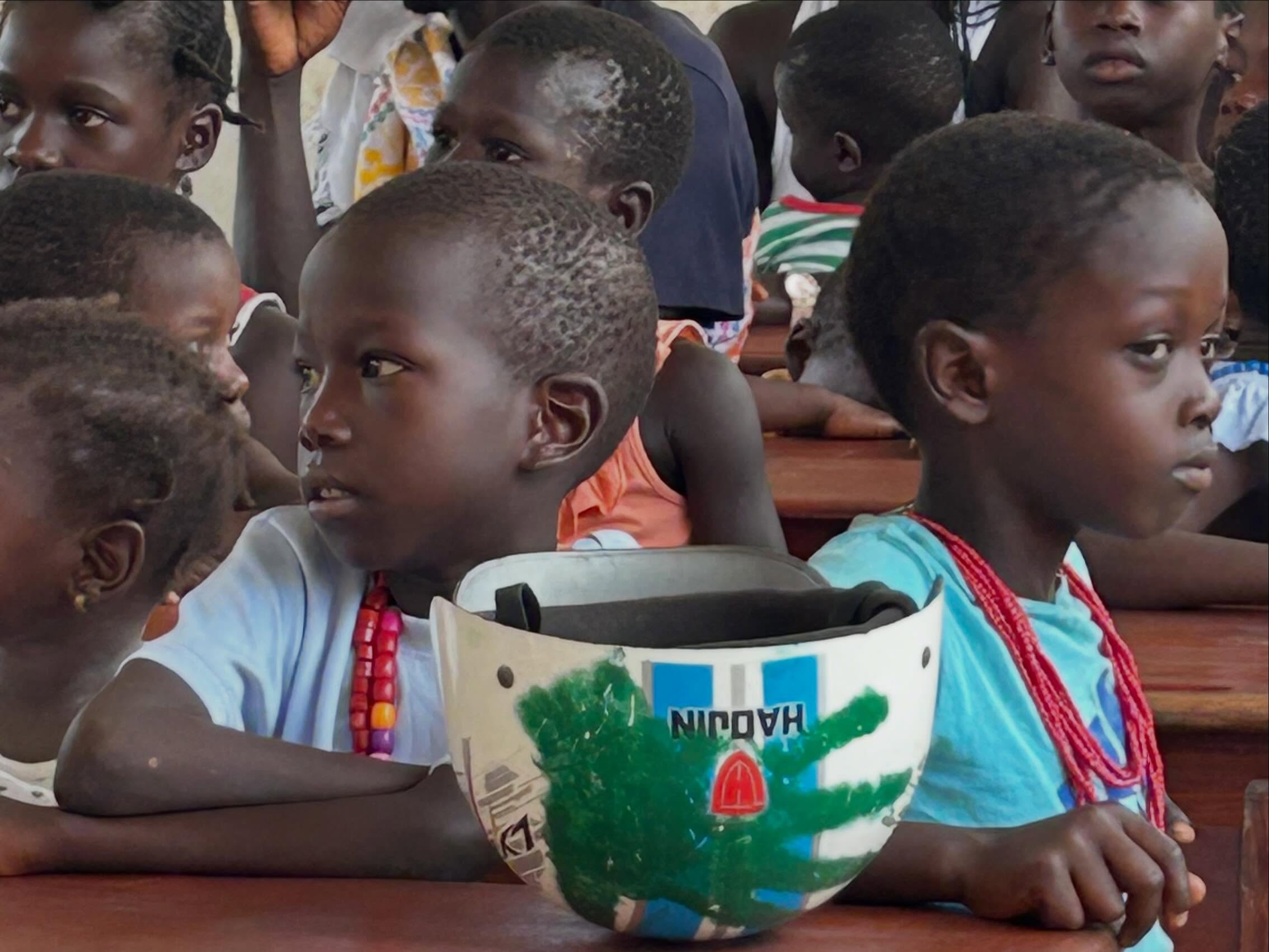 Crianças sentadas em carteiras escolares, numa sala de aula na Guiné-Bissau, com uma tigela pintada à frente de uma delas, ilustrando o ambiente de aprendizagem apoiado pela missão solidária.